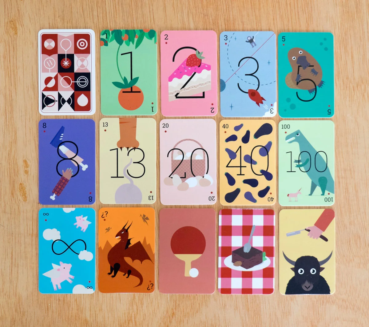 Photo of Redbooth's printed planning poker deck on a wooden table: the patterned card back, then cards 1, 2, 3, 5, 8, 13, 20, 40 and 100, the infinity card, the question mark card, a ping-pong paddle for a break, and two joke cards, a brownie and a yak. Each number is drawn with an illustration, such as a slice of cake for 2, a platypus for 5 and a dinosaur for 100.